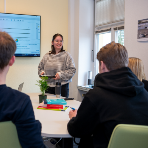 Auszubildende steht vor einer Gruppe von Menschen in einem Besprechungsraum und lacht